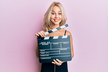 Beautiful caucasian blonde girl holding video film clapboard smiling with a happy and cool smile on...