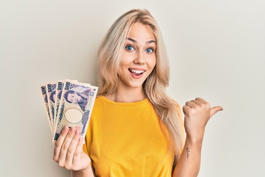 Beautiful caucasian blonde girl holding 5000 japanese yen banknotes pointing thumb up to the side smiling happy with open mouth