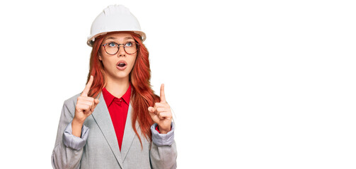 Young redhead woman wearing architect hardhat amazed and surprised looking up and pointing with fingers and raised arms.