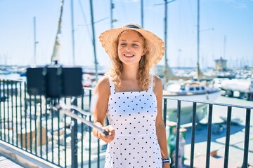 Young beautiful caucasian woman with blond hair smiling happy outdoors taking a selfie picture using selfie stick