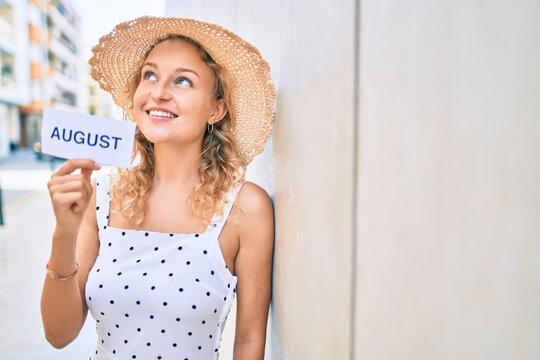 Young beautiful caucasian woman with blond hair smiling happy outdoors on a summer day holding August word
