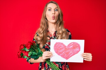 Young caucasian woman with blond hair holding heart draw making fish face with mouth and squinting eyes, crazy and comical.