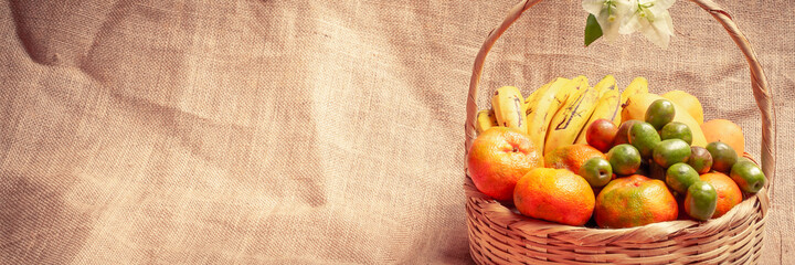 Tropiocal fruits in a wooden basket. Mandarins, tropical plums, bananas and mango, jute background. PAnoramic image with copy space.