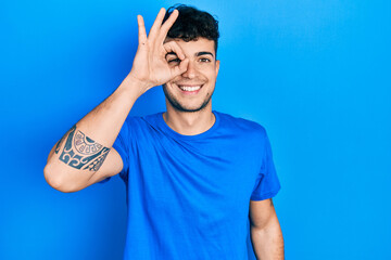 Young hispanic man wearing casual blue t shirt doing ok gesture with hand smiling, eye looking through fingers with happy face.