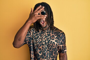 African american man with braids wearing animal print shirt and sunglasses peeking in shock covering face and eyes with hand, looking through fingers afraid
