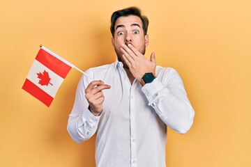 Handsome caucasian man with beard holding canada flag covering mouth with hand, shocked and afraid...