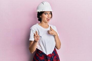 Young caucasian woman wearing hardhat pointing fingers to camera with happy and funny face. good energy and vibes.