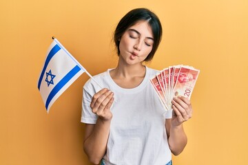 Young caucasian woman holding israel flag and shekels banknotes making fish face with mouth and squinting eyes, crazy and comical.