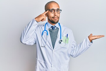 Hispanic adult man wearing doctor uniform and stethoscope confused and annoyed with open palm showing copy space and pointing finger to forehead. think about it.