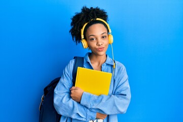 Obraz premium Young african american girl wearing student backpack and headphones holding book looking at the camera blowing a kiss being lovely and sexy. love expression.