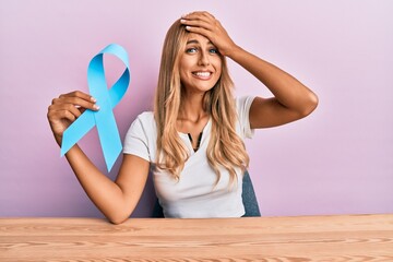 Beautiful blonde young woman holding blue ribbon stressed and frustrated with hand on head,...