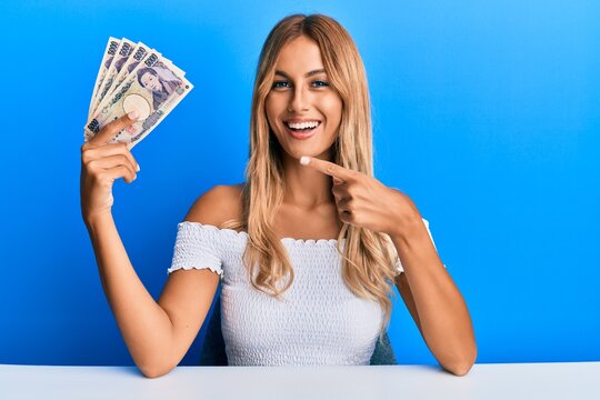 Beautiful blonde young woman holding 5000 japanese yen banknotes smiling happy pointing with hand and finger