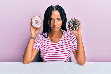 Beautiful hispanic woman holding tasty colorful doughnuts skeptic and nervous, frowning upset...