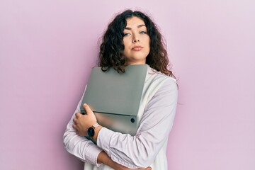 Young brunette woman with curly hair hugging laptop with love relaxed with serious expression on...