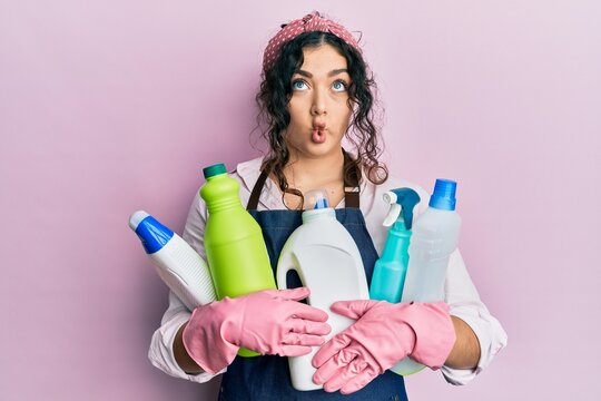 Young brunette woman with curly hair wearing cleaner apron holding cleaning products making fish face with mouth and squinting eyes, crazy and comical.