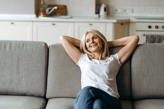 Happy Mature Caucasian Woman Is Relaxing On The Couch In Living Room At Home. Middle Aged Woman Enjoying Weekend Or Leisure From Homework Sitting On Sofa, Looks Away And Smiling