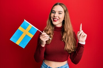 Young blonde woman holding sweden flag smiling with an idea or question pointing finger with happy face, number one