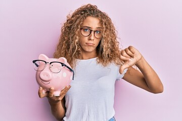 Beautiful caucasian teenager girl holding piggy bank with glasses with angry face, negative sign...