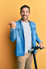 Young handsome man standing on modern scooter over yellow background pointing thumb up to the side...