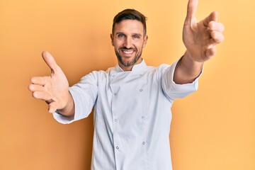 Handsome man with beard wearing professional cook uniform looking at the camera smiling with open...