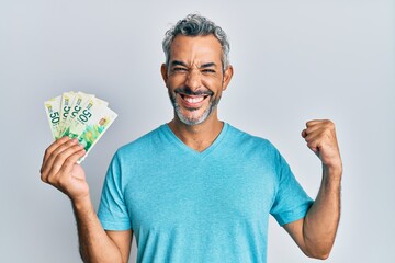 Middle age grey-haired man holding israel shekels banknotes screaming proud, celebrating victory...