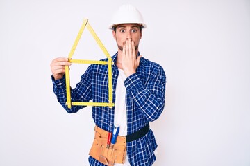 Young handsome man wearing architect hardhat holding build project covering mouth with hand,...