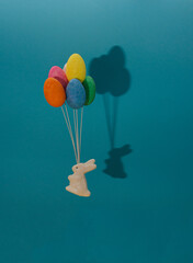 A white rabbit flies with balloons of colorful eggs. Blue background with shadow of bunny and easter eggs.