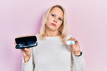Beautiful caucasian blonde woman holding glasses and contact lenses relaxed with serious expression...