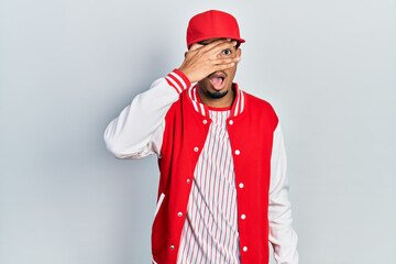 Young african american man wearing baseball uniform peeking in shock covering face and eyes with hand, looking through fingers with embarrassed expression.