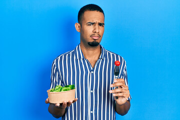Young african american man eating salad clueless and confused expression. doubt concept.