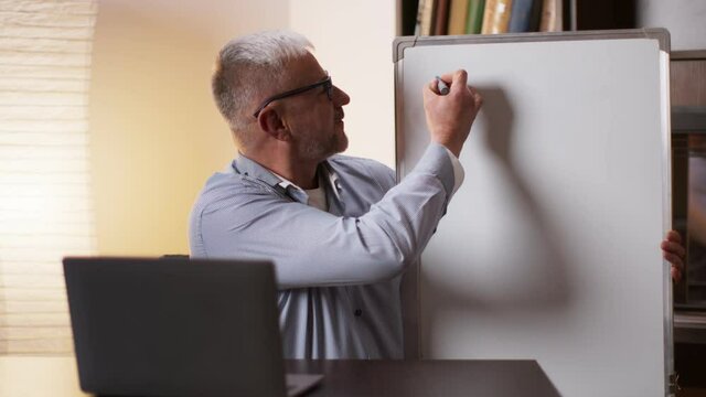Middle aged math professor writing on whiteboard, showing and explaining formulas to students online via video call