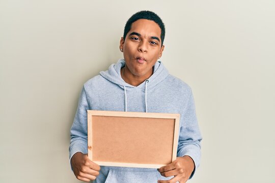 Young handsome hispanic man holding empty corkboard making fish face with mouth and squinting eyes, crazy and comical.
