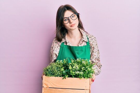 Young beautiful caucasian girl wearing gardener apron holding wooden plant pot relaxed with serious expression on face. simple and natural looking at the camera.