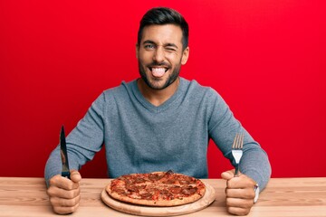 Handsome hispanic man eating tasty pepperoni pizza sticking tongue out happy with funny expression.