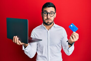 Young hispanic man wearing business style holding laptop and credit card puffing cheeks with funny face. mouth inflated with air, catching air.