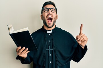Young hispanic priest man holding bible with finger up angry and mad screaming frustrated and furious, shouting with anger looking up.
