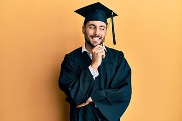 Young hispanic man wearing graduation cap and ceremony robe with hand on chin thinking about question, pensive expression. smiling with thoughtful face. doubt concept.