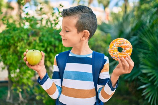 Adorable caucasian student boy smiling happy holding donut and bottle of water at the park.
