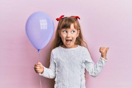 Little caucasian girl kid holding purple balloon pointing thumb up to the side smiling happy with open mouth