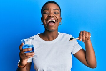 Young african american woman holding glass of mouthwash and toothbrush for fresh breath smiling and...