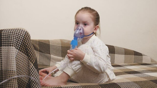 Treatment of a child at home for the flu and colds of the lungs. Kid will be inhaled with a nebulizer while sitting on the sofa in the children's room. A little girl breathes into a mask with medicine