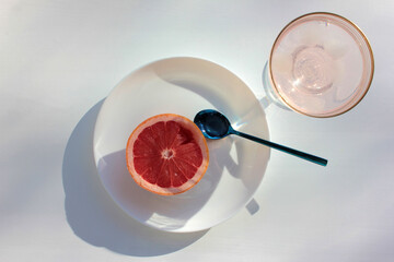 Wide glass of pink champagne cocktail and half of ripe grapefruit on plate and spoon on white background. Flat lay food composition with bright citrus fruit on table. Hard light, shadow. Top view

