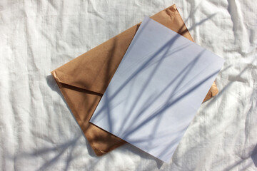 Brown kraft envelope and white blank paper card on bed crumpled texture with palm leaves shadows. Flat lay composition with mock-up invitation. Top view, copy space. Hard light. Summer still life