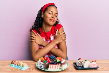 Young african american girl wearing baker uniform sitting on the table with sweets hugging oneself happy and positive, smiling confident. self love and self care