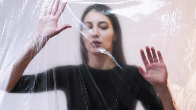 Female rights. Defocused portrait. Human trafficking. Slavery abuse. Scared disturbed woman hostage isolated trapped behind transparent wrinkled polyethylene film on light background out of focus.