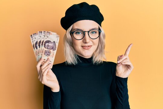 Young blonde girl holding mexican pesos smiling happy pointing with hand and finger to the side