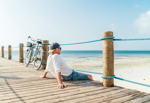 Portrait of relaxing man dressed in light summer clothes and sunglasses sitting and enjoying time and beach view on wooden pier.Careless vacation in tropical countries concept image. Zanzibar,Tanzania