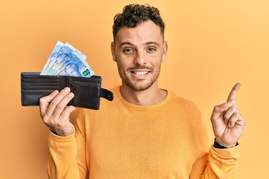 Young hispanic man holding wallet with south african rand banknotes smiling happy pointing with hand and finger to the side