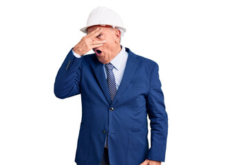 Senior handsome grey-haired man wearing suit and architect hardhat peeking in shock covering face and eyes with hand, looking through fingers with embarrassed expression.