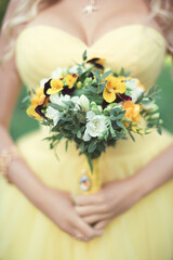 Obraz na płótnie Canvas A delicate and elegant bride's bouquet of yellow-purple pansies, white and orange freesia and greenery with a vintage brooch on the ribbon. Details of an exquisite yellow country wedding ceremony.